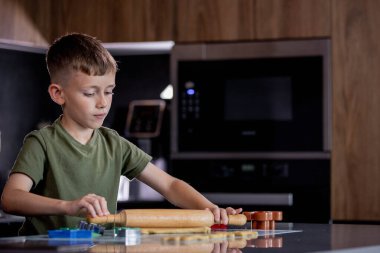 Küçük çocuk masaya merdaneyle zencefilli ekmek hamuru yuvarlıyor.