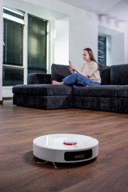 Bir kadın telefonuna bakarken bir robot süpürge odayı temizliyor..