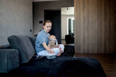 Anne karanlık bir koltukta oturup çocuğunu tutuyor ve evde astım spreyini kullanmalarına yardım ediyor.