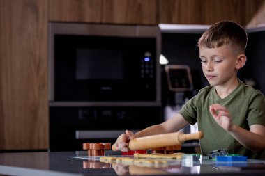 Küçük çocuk masaya merdaneyle zencefilli ekmek hamuru yuvarlıyor.