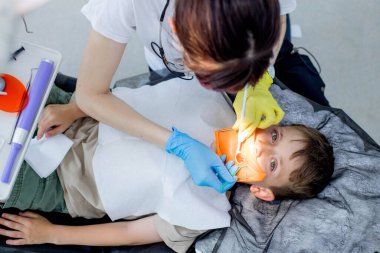 Çocuk dişçiliği. Dişçide ilk muayene. Doktor dişlerini tedavi ederken ağzı açık yakışıklı bir çocuk yan bakıyor.
