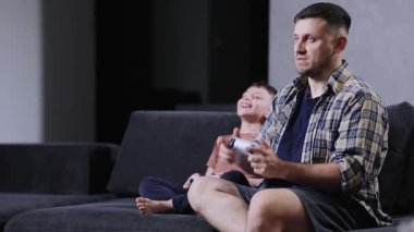 Baba ve küçük oğlu oturma odasında kanepede oturup evde video oyunları oynuyorlar.