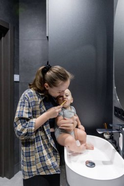 Bir kadın, banyoda temiz ve düzenli bir şekilde bir bebeği kollarında tutarken dişlerini özenle fırçalıyor.