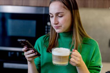 Genç bir kadın mutfakta telefona bakıyor ve kahve makinesinde kahve yapıyor..