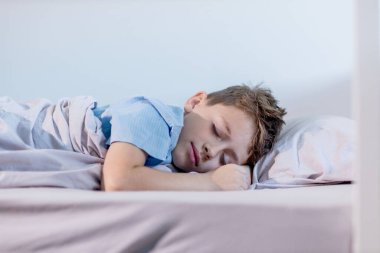 Mavi tişörtlü genç bir çocuk rahat yatağında uyur yumuşak battaniyelerle sarılı ve rahat çarşaflarla çevrili.