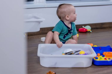 Genç bir bebek, temiz beyaz bir çöp kutusunda saklanan renkli oyuncaklarla mutlu bir şekilde oynuyor. Keşif ve etkileşimlerinden keyif alıyor.