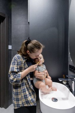 Bir kadın, banyoda temiz ve düzenli bir şekilde bir bebeği kollarında tutarken dişlerini özenle fırçalıyor.