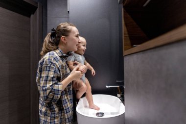 Bir kadın küçük bir bebeği nazikçe kollarında tutarak banyoda durarak sıcaklık ve özenle dolu bir sahne yaratıyor..