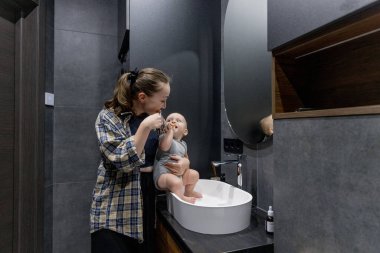 Bir kadın, banyoda temiz ve düzenli bir şekilde bir bebeği kollarında tutarken dişlerini özenle fırçalıyor.