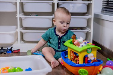 Küçük bir bebek rahat bir şekilde yumuşak zeminde oturuyor, etrafında çeşitli renkli oyuncaklarla neşeyle oynuyor.