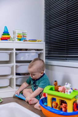 Küçük bir bebek rahat bir şekilde yumuşak zeminde oturuyor, etrafında çeşitli renkli oyuncaklarla neşeyle oynuyor.