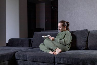 Bir kadın rahat bir şekilde rahat bir kanepede oturuyor, keyifli bir öğleden sonra ilginç bir kitap okumaya dalmış.