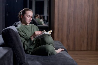 Bir kadın rahat bir şekilde rahat bir kanepede oturuyor, keyifli bir öğleden sonra ilginç bir kitap okumaya dalmış.