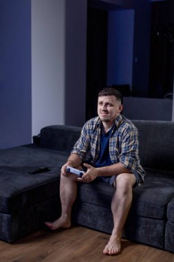 Bir adam elinde bir video oyun kumandası tutarken koltukta rahatça oturur ve oynadığı oyuna dalmıştır.