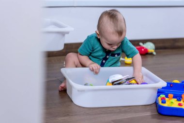 Genç bir bebek, temiz beyaz bir çöp kutusunda saklanan renkli oyuncaklarla mutlu bir şekilde oynuyor. Keşif ve etkileşimlerinden keyif alıyor.