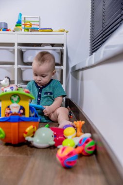 Küçük bir bebek rahat bir şekilde yumuşak zeminde oturuyor, etrafında çeşitli renkli oyuncaklarla neşeyle oynuyor.
