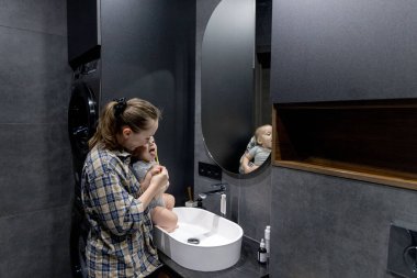 Bir kadın, banyoda temiz ve düzenli bir şekilde bir bebeği kollarında tutarken dişlerini özenle fırçalıyor.