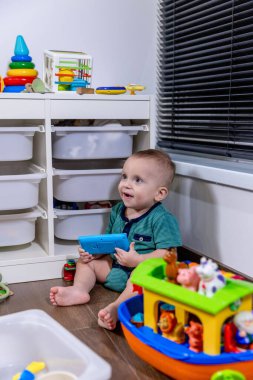 Küçük bir bebek rahat bir şekilde yumuşak zeminde oturuyor, etrafında çeşitli renkli oyuncaklarla neşeyle oynuyor.