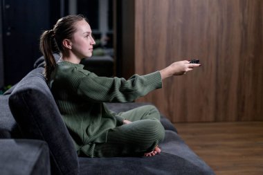 Rahat bir şekilde rahat bir koltuğa yerleştirilmiş bir kadın elinde uzaktan kumandayla kanalı ya da sesi değiştirmeye hazırdır.