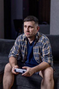 Bir adam elinde bir video oyun kumandası tutarken koltukta rahatça oturur ve oynadığı oyuna dalmıştır.