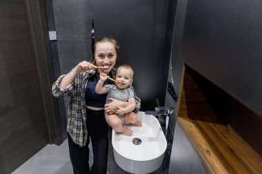 Bir kadın, banyoda temiz ve düzenli bir şekilde bir bebeği kollarında tutarken dişlerini özenle fırçalıyor.