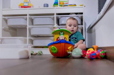 Küçük bir bebek rahat bir şekilde yumuşak zeminde oturuyor, etrafında çeşitli renkli oyuncaklarla neşeyle oynuyor.