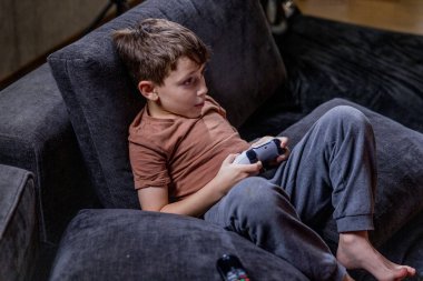 Oğlan evdeki kanepede oturup oyun oynuyor.