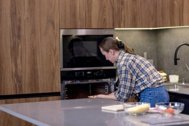 Bir kadın, modern mutfağındaki modern bir fırına nefis bir yemek koyar. Çağdaş mutfak yetenekleri sergiler..