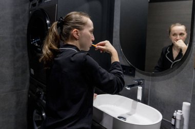Rahat bir pijama giymiş bir kadın modern bir banyoda dişlerini fırçalıyor ve günlük bakım rutininin tadını çıkarıyor..