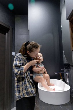 Bir kadın, banyoda temiz ve düzenli bir şekilde bir bebeği kollarında tutarken dişlerini özenle fırçalıyor.