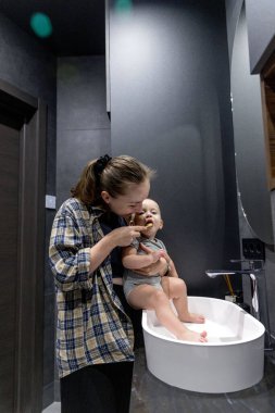 Bir kadın, banyoda temiz ve düzenli bir şekilde bir bebeği kollarında tutarken dişlerini özenle fırçalıyor.