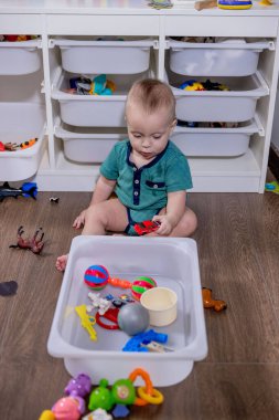 Genç bir bebek, temiz beyaz bir çöp kutusunda saklanan renkli oyuncaklarla mutlu bir şekilde oynuyor. Keşif ve etkileşimlerinden keyif alıyor.