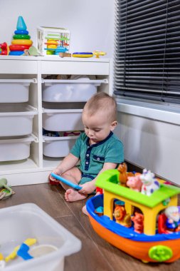 Küçük bir bebek rahat bir şekilde yumuşak zeminde oturuyor, etrafında çeşitli renkli oyuncaklarla neşeyle oynuyor.