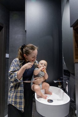 Bir kadın, banyoda temiz ve düzenli bir şekilde bir bebeği kollarında tutarken dişlerini özenle fırçalıyor.