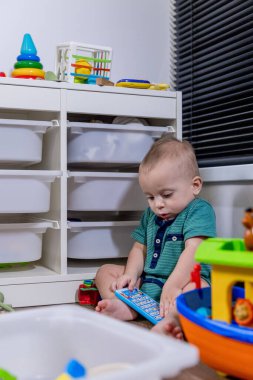 Küçük bir bebek rahat bir şekilde yumuşak zeminde oturuyor, etrafında çeşitli renkli oyuncaklarla neşeyle oynuyor.