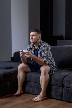 Bir adam elinde bir video oyun kumandası tutarken koltukta rahatça oturur ve oynadığı oyuna dalmıştır.