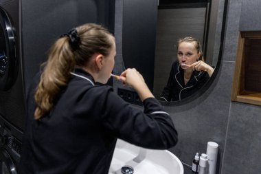 Rahat bir pijama giymiş bir kadın modern bir banyoda dişlerini fırçalıyor ve günlük bakım rutininin tadını çıkarıyor..