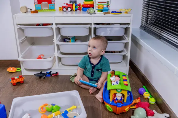 Küçük bir bebek rahat bir şekilde yumuşak zeminde oturuyor, etrafında çeşitli renkli oyuncaklarla neşeyle oynuyor.