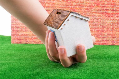 Yeşil çim ile kırmızı tuğla arka planda bir ev modeli tutan el