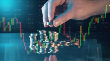 A hand adds coins to a pile with stock charts on the screen. 4K