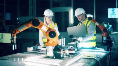 Two engineers in safety wear are observing a robotic arm. 4K