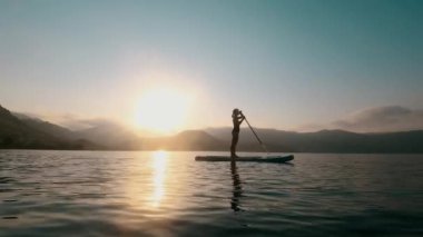 Güneş doğarken bir SUP tahtasındaki kadın parlayan suda kürek çekiyor. 4K
