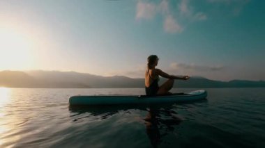 Gün batımında SUP sörf tahtasında yavaş çekim 4K ile yoga yapan tanınmayan bir kadın.