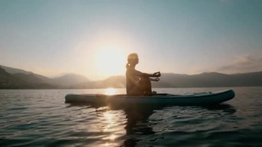 Gün batımında SUP panosunda yoga yapan genç bir kadın. Yavaş çekim gelecek
