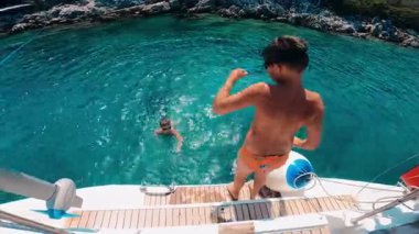 Küçük bir teknede poz veren genç bir çocuk aşağıdaki ışıldayan turkuaz suya atlamaya hevesle hazırlanıyor.