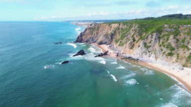 Portekiz, Sintra 'daki Atlantik Okyanusu' nun yüksek kıyısındaki geniş insansız hava aracı panoraması. Parlak güneşli bir gün. Kayaların altındaki sarı kumların üzerinde uzun beyaz dalgalar.