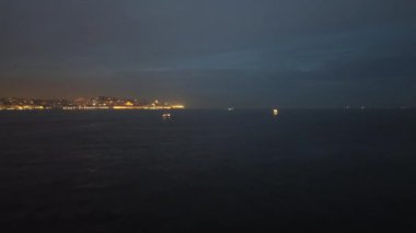 İnsansız hava aracı gece nehrinin üzerinden şehre ve gece ışıklarına doğru alçaktan uçuyor..