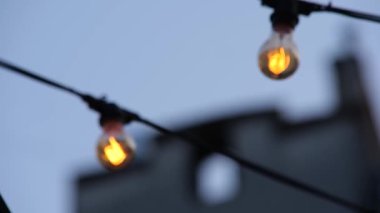 Kablolardan sarkan iki Edison tarzı ampul. Ampullerin içindeki liflerin yumuşak parıltısı solan mavi gökyüzü ile güzel bir tezat oluşturuyor. Eski ya da rahat bir atmosfer.