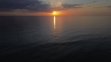 Okyanusun üzerinde sakin bir günbatımı, suyun üzerinde güneşe doğru uçan bir dron. Altın ışık sakin suya güzel bir şekilde yansıyor. Yumuşak bulutlar gökyüzünde sürükleniyor, sakin atmosferi güçlendiriyor..