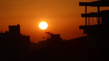 Şehir manzarasında gün doğumu: Turuncu bir gökyüzünde parlak bir sabah güneşi yükseliyor, binaların ve inşaatın karanlık siluetlerini aydınlatıyor. Sabahın erken saatlerinde şehir sakinliği..
